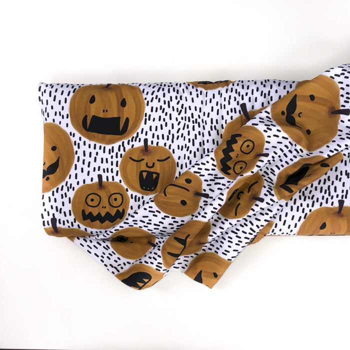 Stock image of mage of Lillestoff Pumpkin Organic cotton jersey fabric on a roll. Fabric is white with little black sticks in the background. There are different sizes orange pumpkins with faces on them.